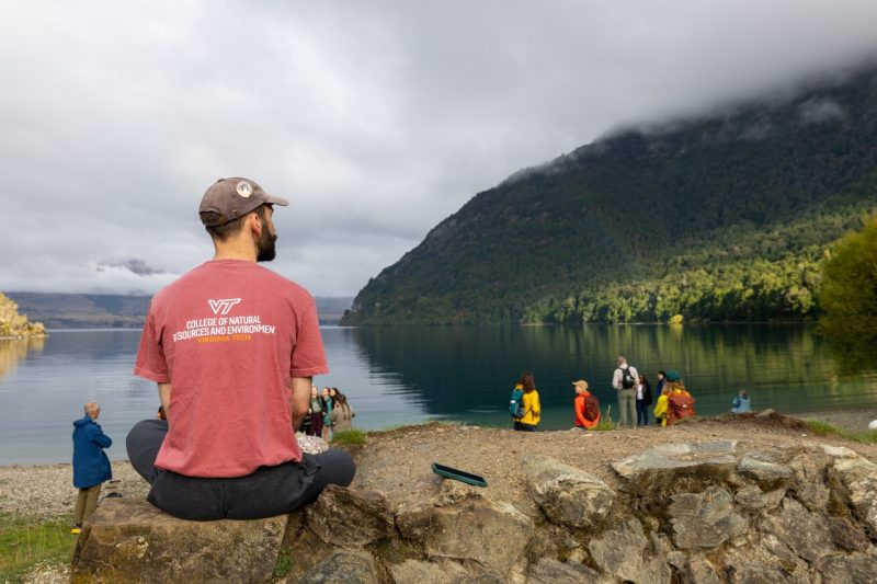 man (in foreground at left) sits on a rock in a virginia tech shirt in front of a lake